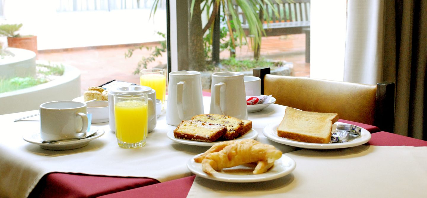 Mesa servida con café, jugo de naranja, tostadas y medialunas frente a la ventana