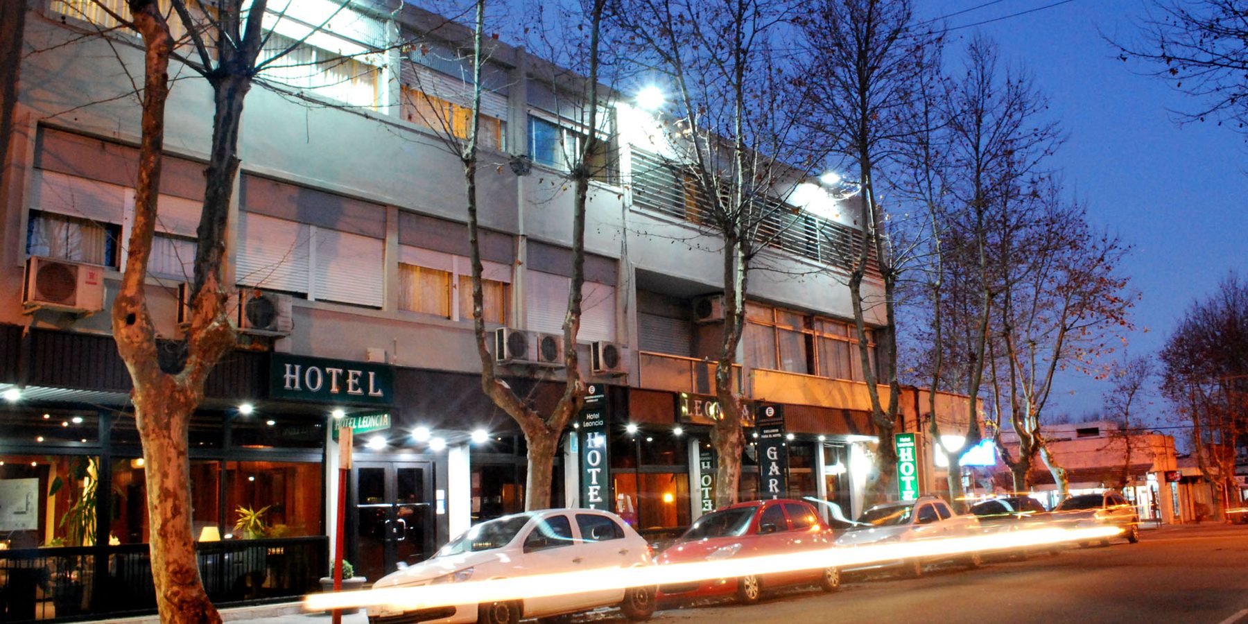 La calle Rivera de noche frente al hotel, con los plátanos y las luces de los autos