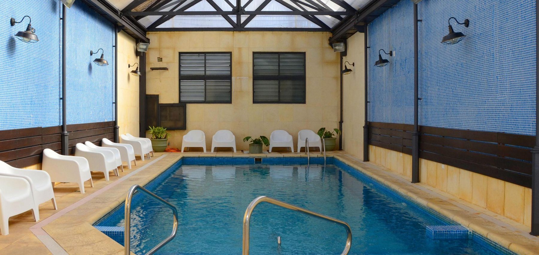 Vista lateral de la piscina climatizada con sillones blancos y farolas de pared