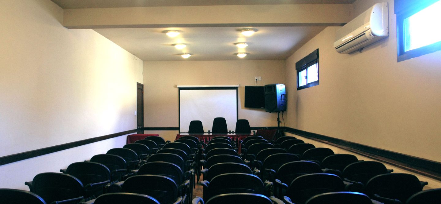 Sala de reuniones con filas de sillas negras, pantalla y televisor